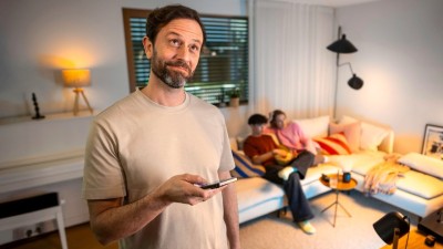 Ein Mann steuert die smarte Beleuchtung mit dem Smartphone, im Hintergrund sitzt die Familie auf dem Sofa.