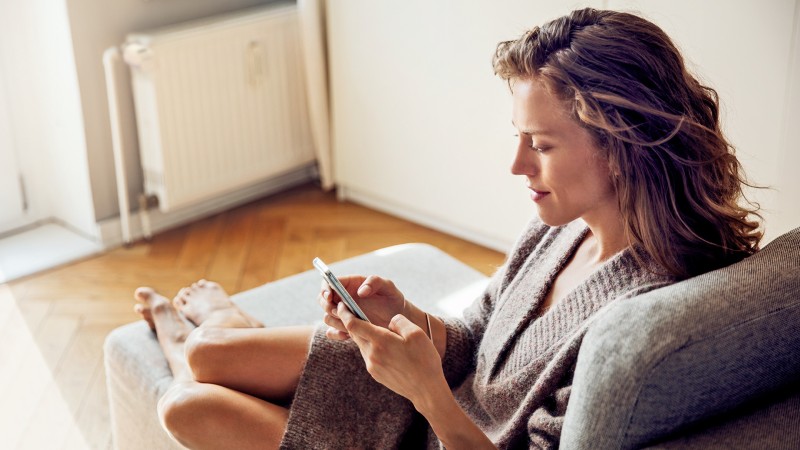 Eine Frau sitzt auf einem Sofa und ist am Handy im Hintergrund eine Heizung