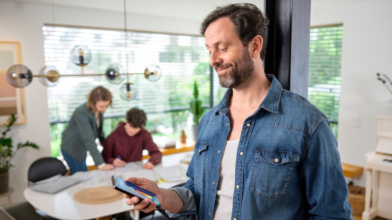 A family man controls the heating with his smartphone while his widget is doing homework with the child in the background.