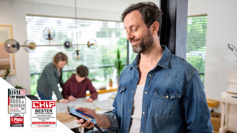 A family man controls the heating with his smartphone while his widget is doing homework with the child in the background.